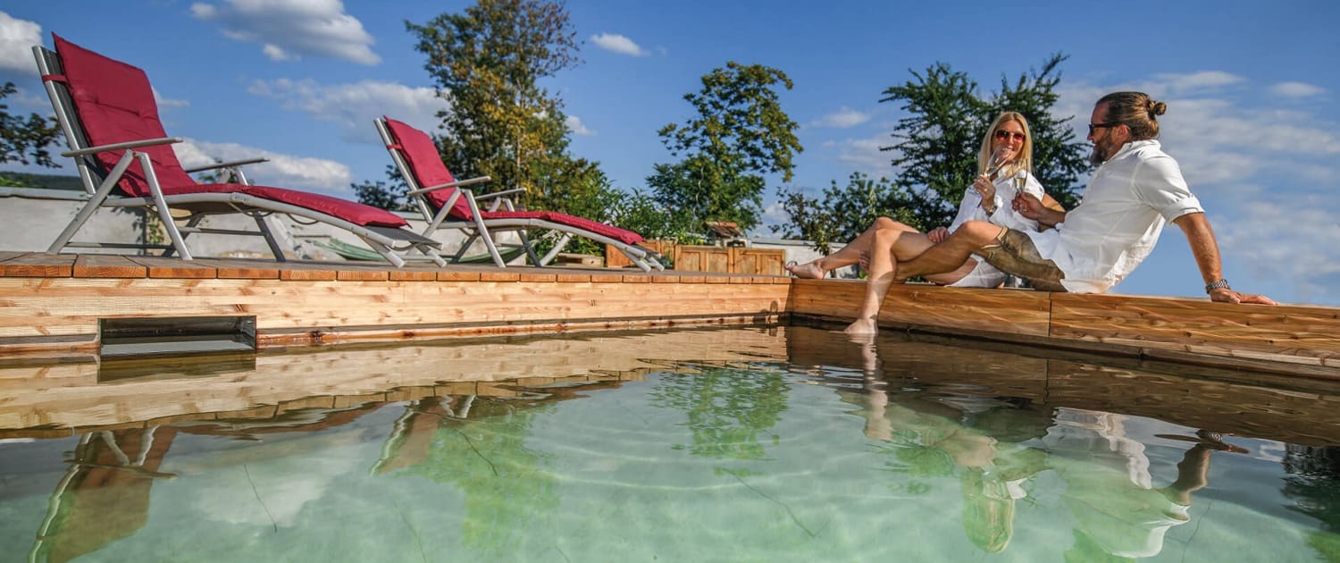 Pärchen trinkt Sekt am Holc Naturpool. Dem Naturpool aus Holz.